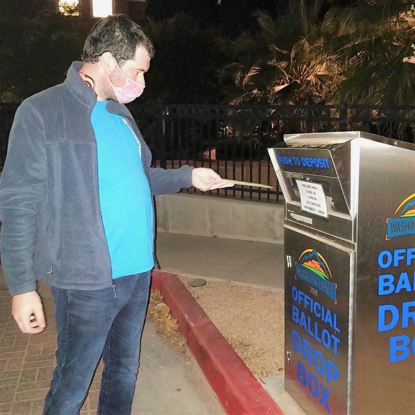 photo of a young adult with a mask on dropping a ballot into an official ballot dropbox.